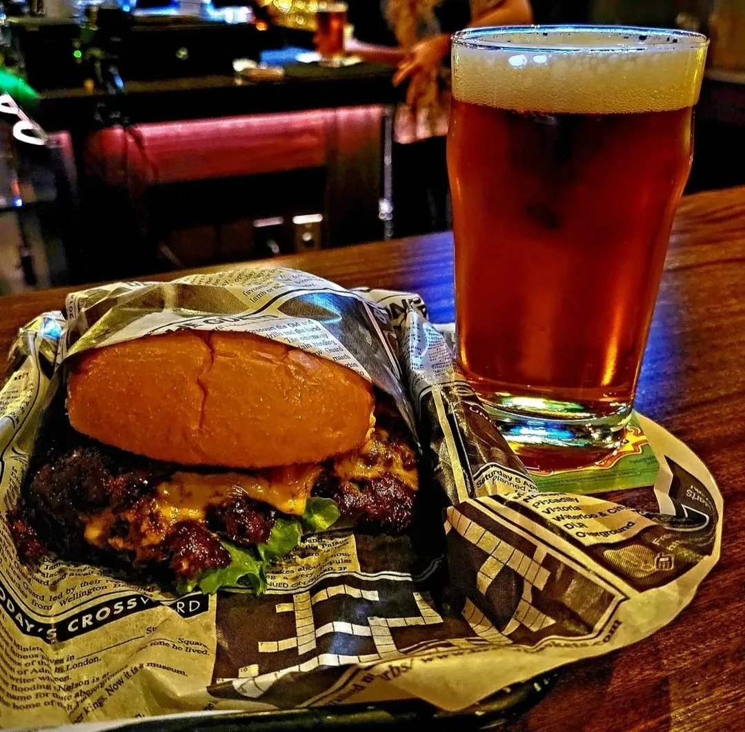 Smash Burger with melted cheese, wrapped in newspaper, next to a beer.